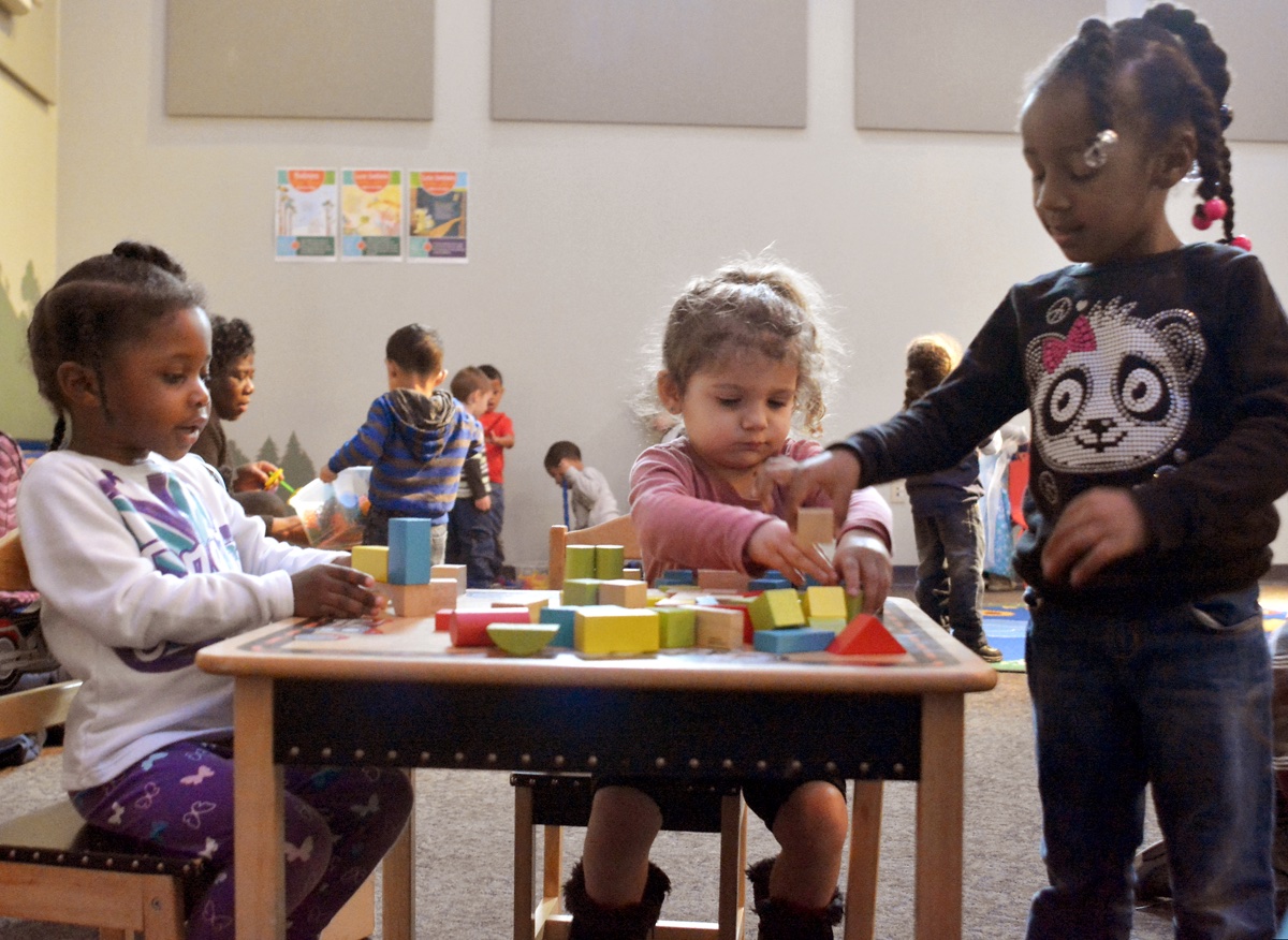 three pre-school age girls play with blocks in a community room