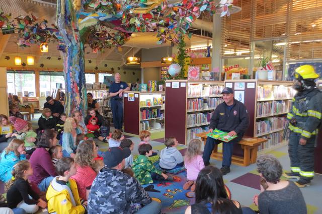Two firefighters reading a book to a group of children in a library.